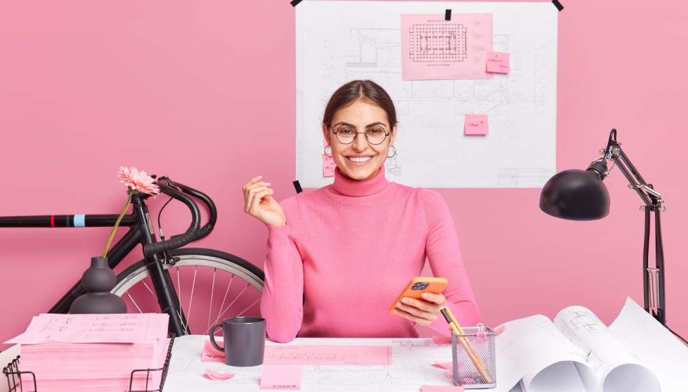 cheerful-female-graphic-designer-holds-modern-smartphone-creats-blueprint-drawing-sketch-poses-desktop-wears-spectacles-turtleneck-sits-coworking-space-successful-architect-woks-project